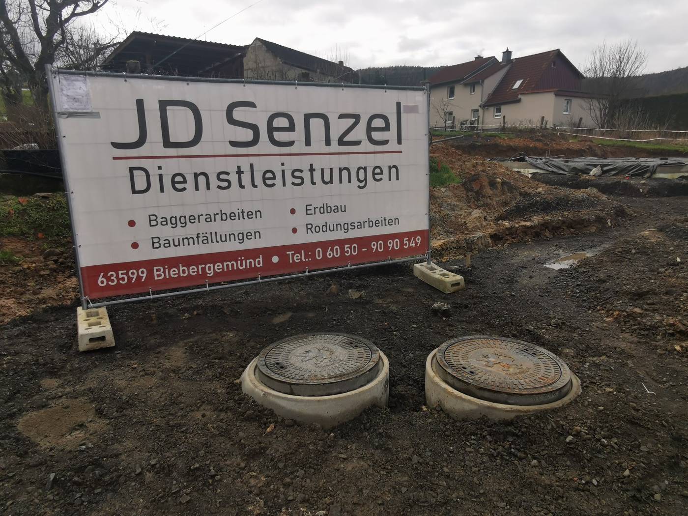 Bauzaunbanner von JD Senzel Dienstleistungen an einer Baustelle mit zwei Revisionsschächten